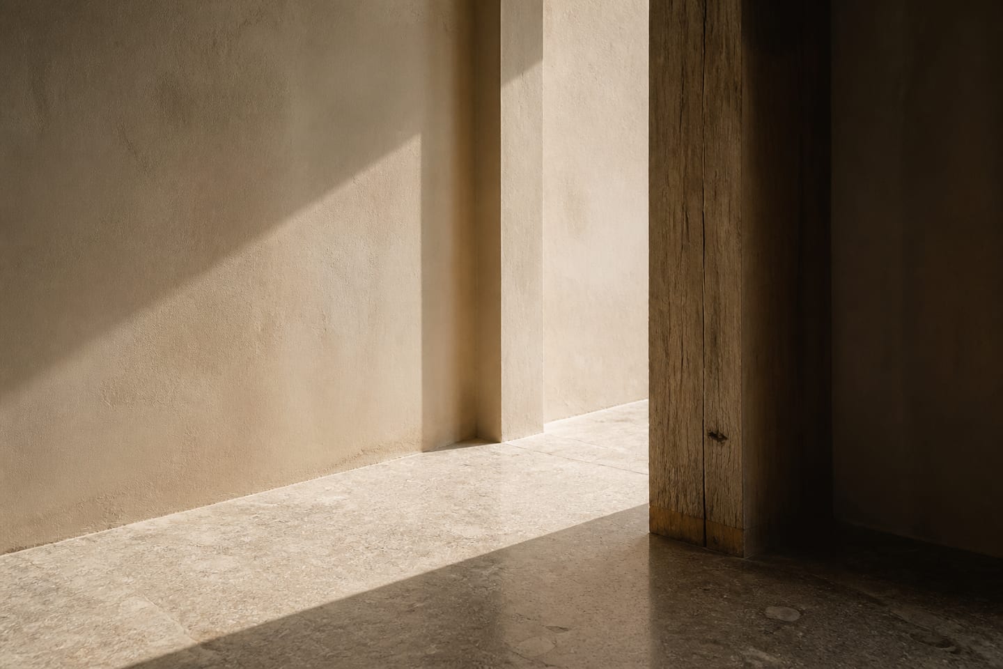 Raking sunlight crossing lime plaster, weathered timber, and a stone floor